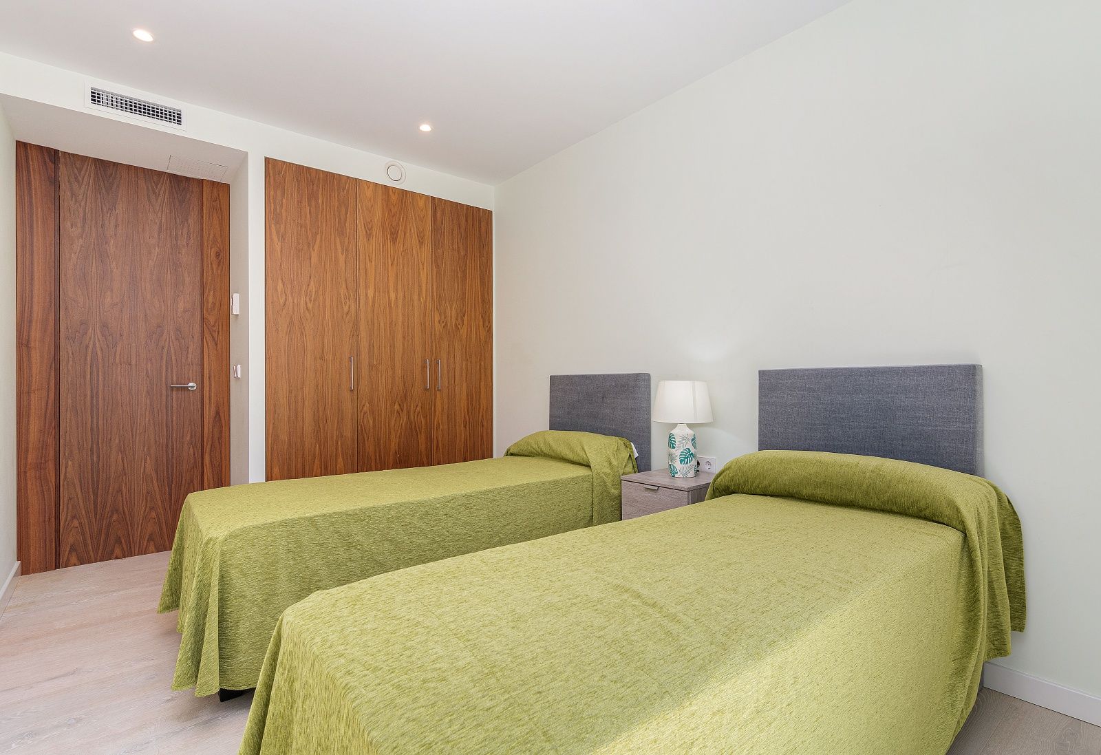 Dormitorio moderno con dos camas individuales, armarios empotrados de madera natural, aire acondicionado por conductos y suelos de tono claro.