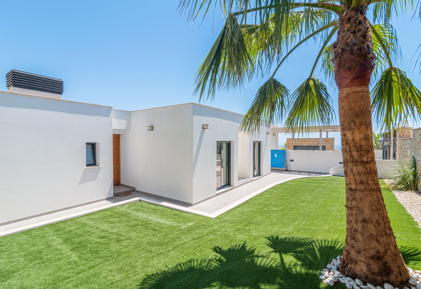 Exterior de villa moderna con fachada blanca, jardín de césped artificial y palmera. Se aprecia la puerta de entrada de madera y amplios ventanales bajo un cielo despejado.