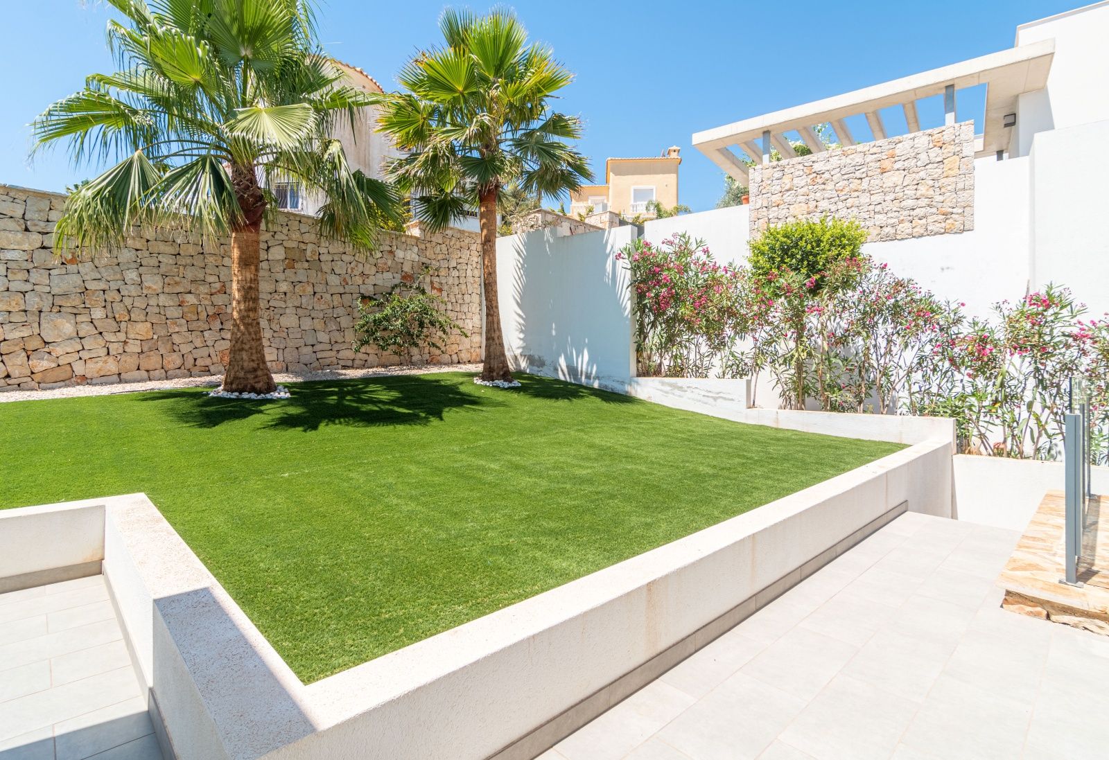 Jardín de diseño con césped artificial, palmeras y muros de piedra seca natural que delimitan la propiedad bajo un cielo despejado.