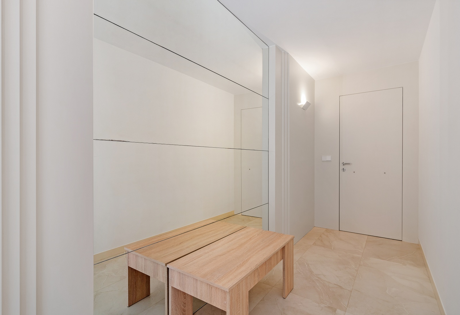 Pasillo de entrada con gran espejo de pared, banco de madera clara y suelos de gres porcelánico en tonos crema. Diseño minimalista con puerta blanca e iluminación de pared.
