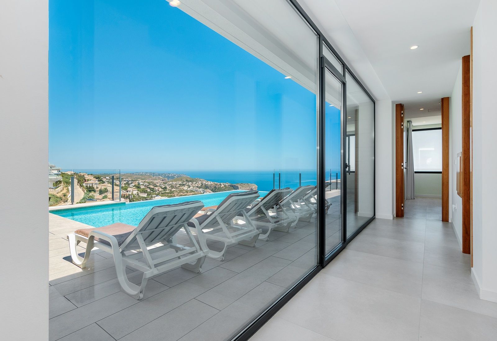 Pasillo interior con grandes ventanales que ofrecen vistas panorámicas al mar y acceso directo a la terraza con piscina infinita y tumbonas.
