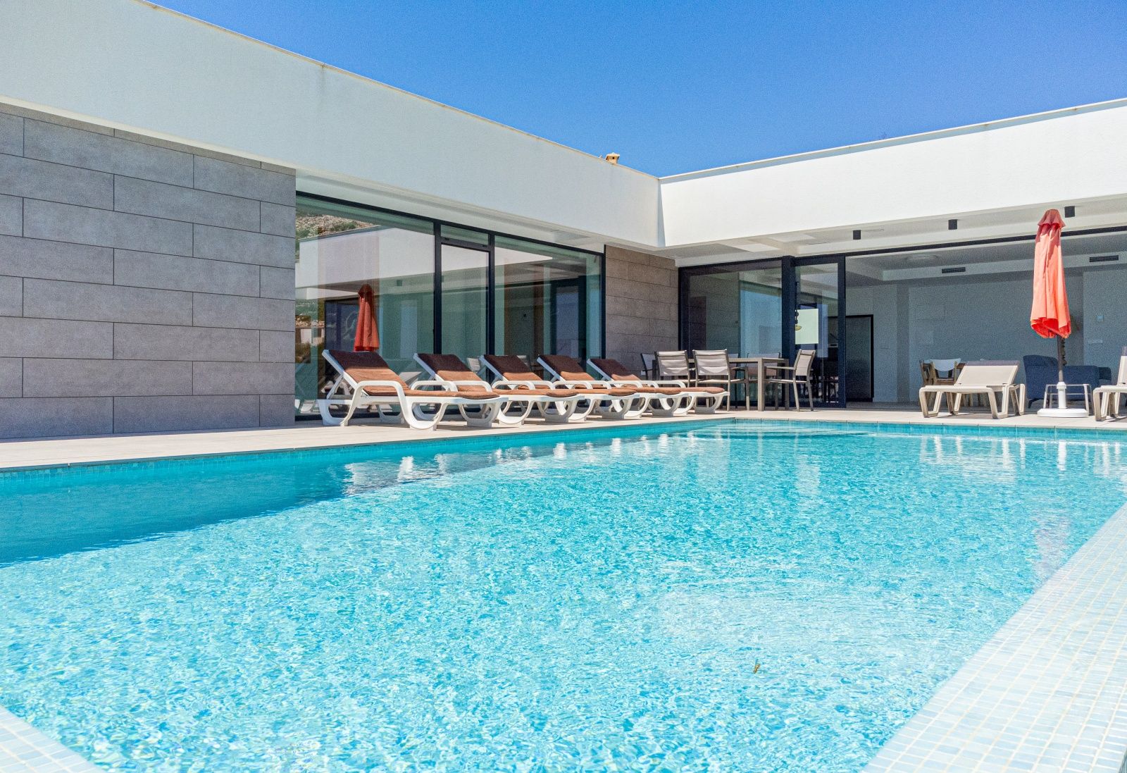 Piscina privada rodeada de una amplia terraza con tumbonas, zona de comedor exterior y fachada moderna con grandes ventanales de cristal bajo un cielo despejado.