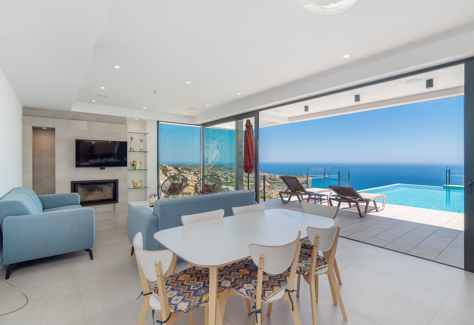 Salón comedor de concepto abierto con grandes ventanales, chimenea integrada y acceso directo a la terraza con piscina infinita y vistas al mar.