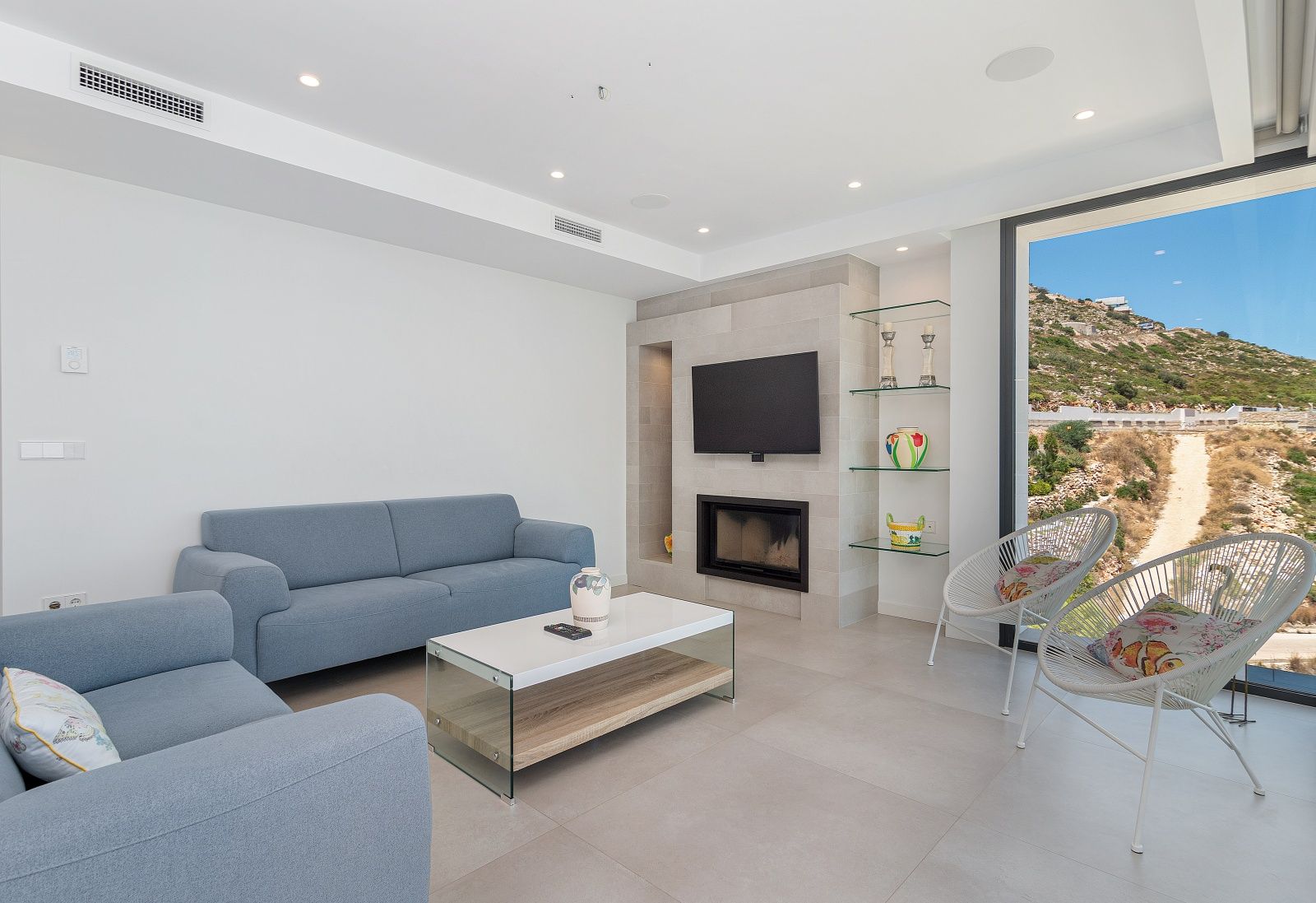 Salón moderno con sofás azules, chimenea integrada en pared de piedra y amplios ventanales con vistas a la montaña. Incluye iluminación empotrada y aire acondicionado.