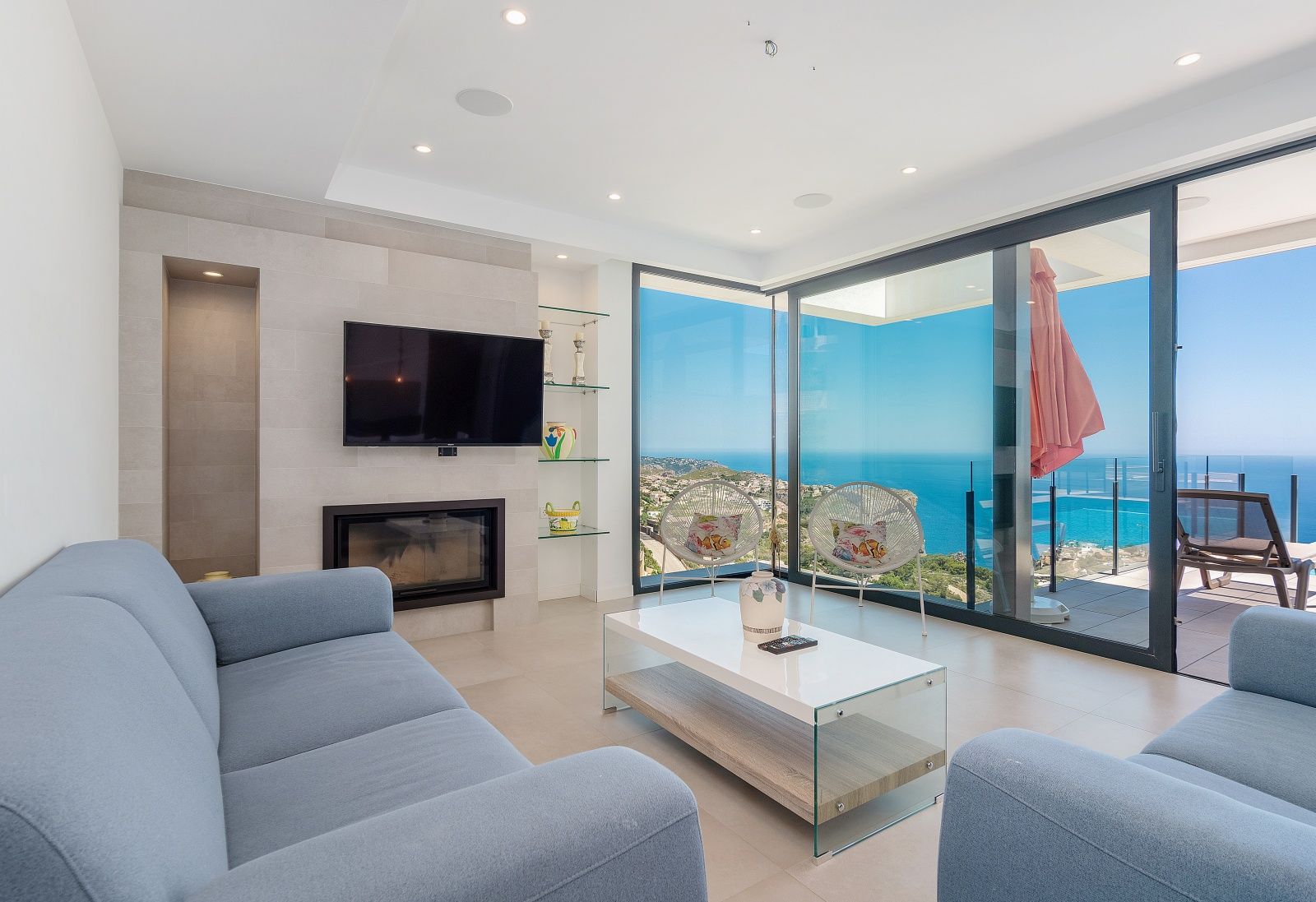 Salón moderno con sofás azules, chimenea integrada en pared de piedra y amplios ventanales con vistas panorámicas al mar Mediterráneo y la costa.