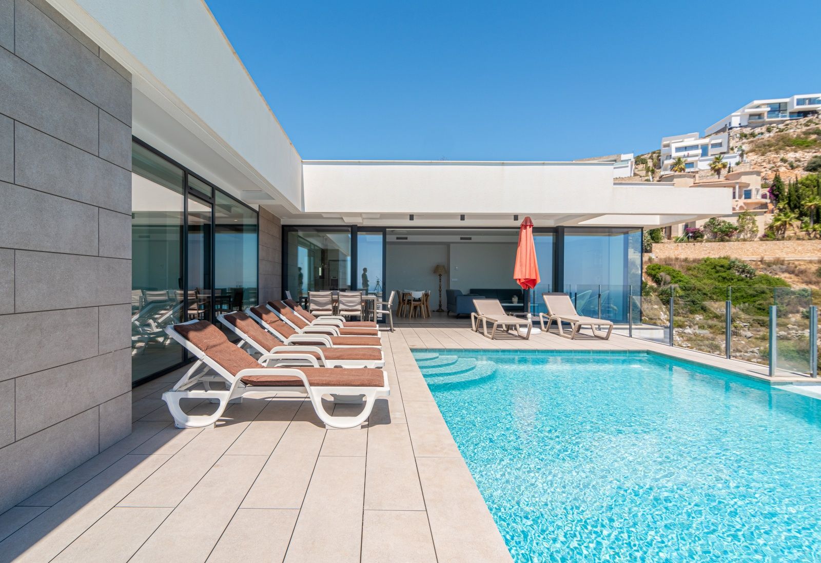 Terraza principal con piscina infinita, hamacas y zona de comedor exterior. Destaca la arquitectura moderna con grandes ventanales y revestimiento de piedra gris.