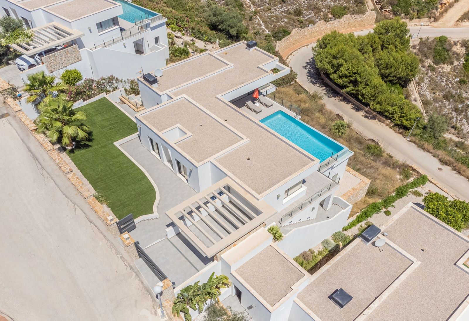 Vista aérea de la villa con arquitectura de líneas rectas, piscina infinita, cubiertas planas de grava y zona ajardinada con césped artificial.
