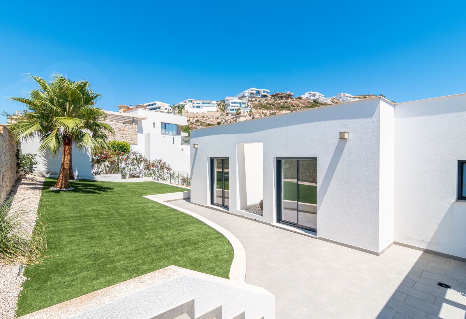 Vista exterior de la villa con fachada blanca minimalista, jardín de césped artificial, una palmera y ventanales con marcos oscuros.