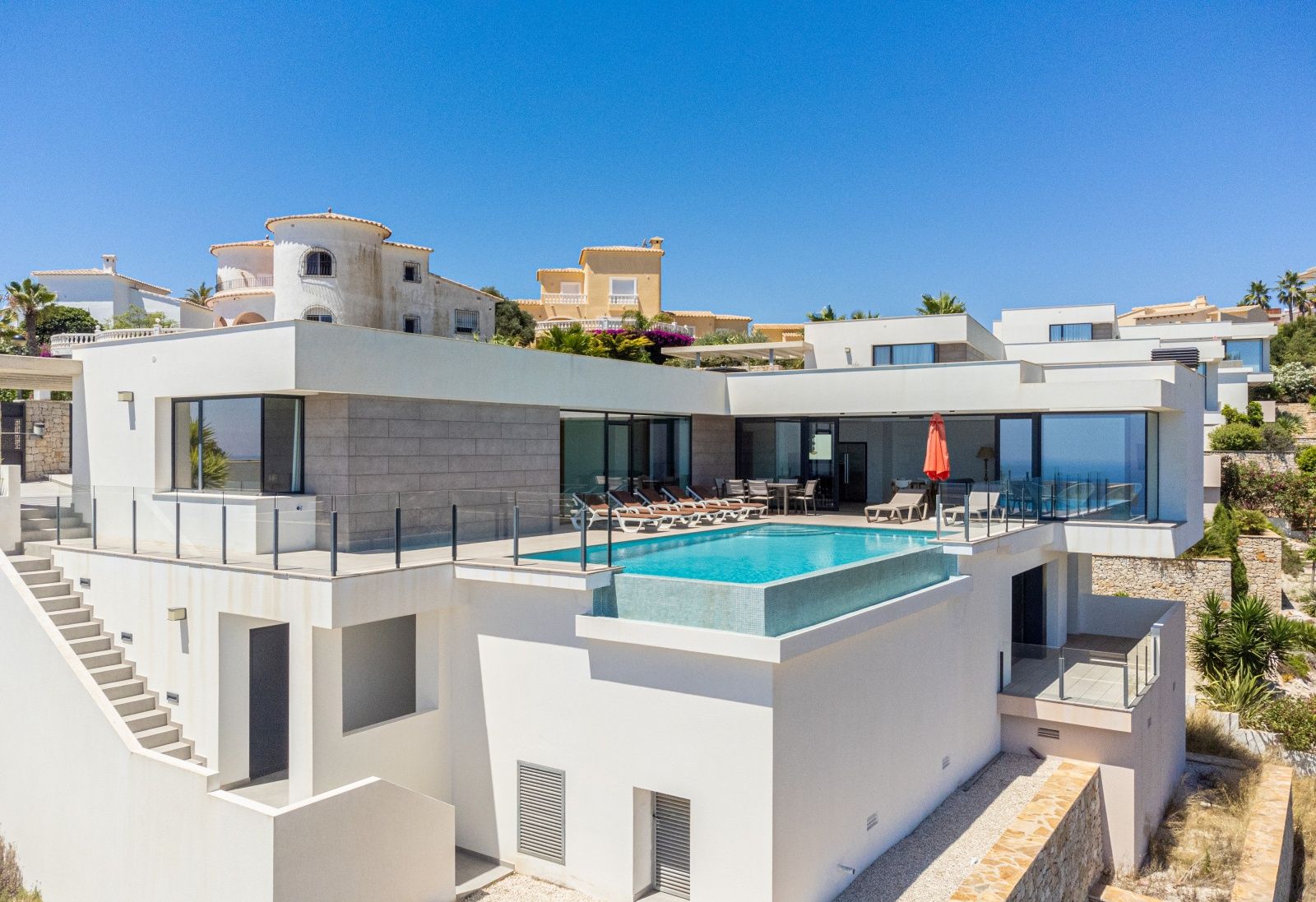 Vista exterior de la villa moderna con piscina infinita, amplia terraza con tumbonas y fachada blanca con detalles en piedra gris.