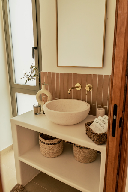 Baño moderno con lavabo de piedra sobre encimera de obra, grifería empotrada dorada y azulejos verticales en tonos tierra. Incluye estante inferior con cestas de mimbre.