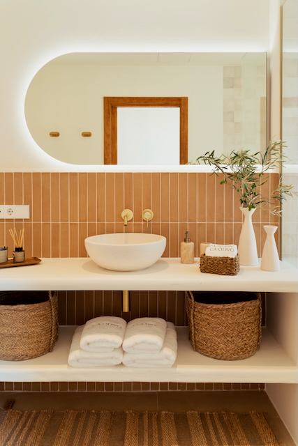 Baño moderno con lavabo sobre encimera, grifería empotrada en latón y azulejos verticales color terracota. Incluye un espejo ovalado retroiluminado y estantes con cestas de mimbre.