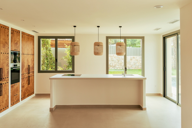 Cocina de concepto abierto con isla central blanca, lámparas de fibra natural y armarios de madera con herrajes. Los ventanales conectan el interior con el jardín y la piscina.