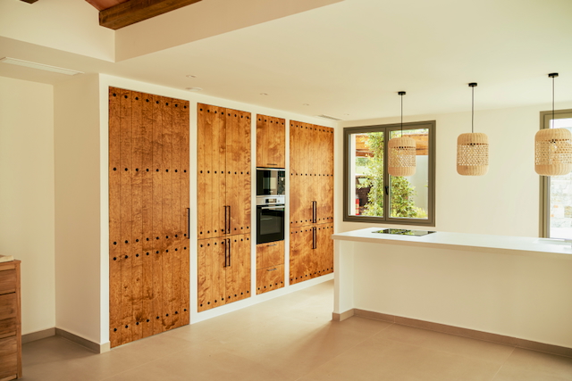 Cocina de planta abierta con armarios de madera rústica y electrodomésticos integrados. Incluye una isla blanca con placa de inducción y lámparas colgantes de mimbre.