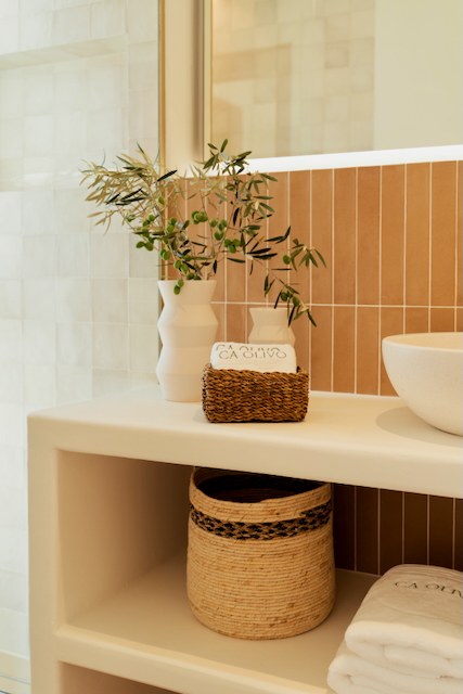 Detalle de baño con encimera de obra blanca, azulejos verticales en tono tierra, lavabo sobre encimera y cestas de fibras naturales bajo el espejo.