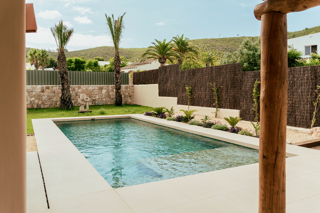 Piscina rectangular con borde de gres claro, integrada en un jardín con palmeras, muros de piedra y vistas a las colinas circundantes.