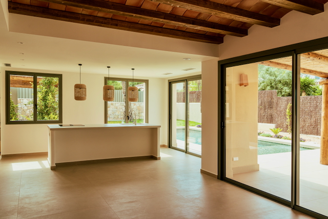 Salón-comedor diáfano con techos de vigas de madera, cocina con isla y acceso directo a la piscina mediante amplios ventanales correderos.