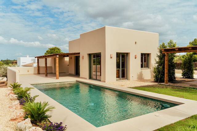 Vista exterior de la villa con piscina rectangular, fachada minimalista en tonos arena y porche con vigas de madera rústica sobre terraza pavimentada.