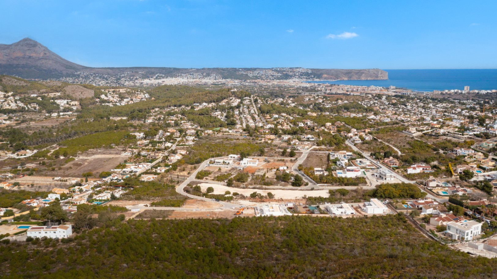 Vista aérea de Jávea con chalets blancos y vegetación, bajo un cielo azul despejado.