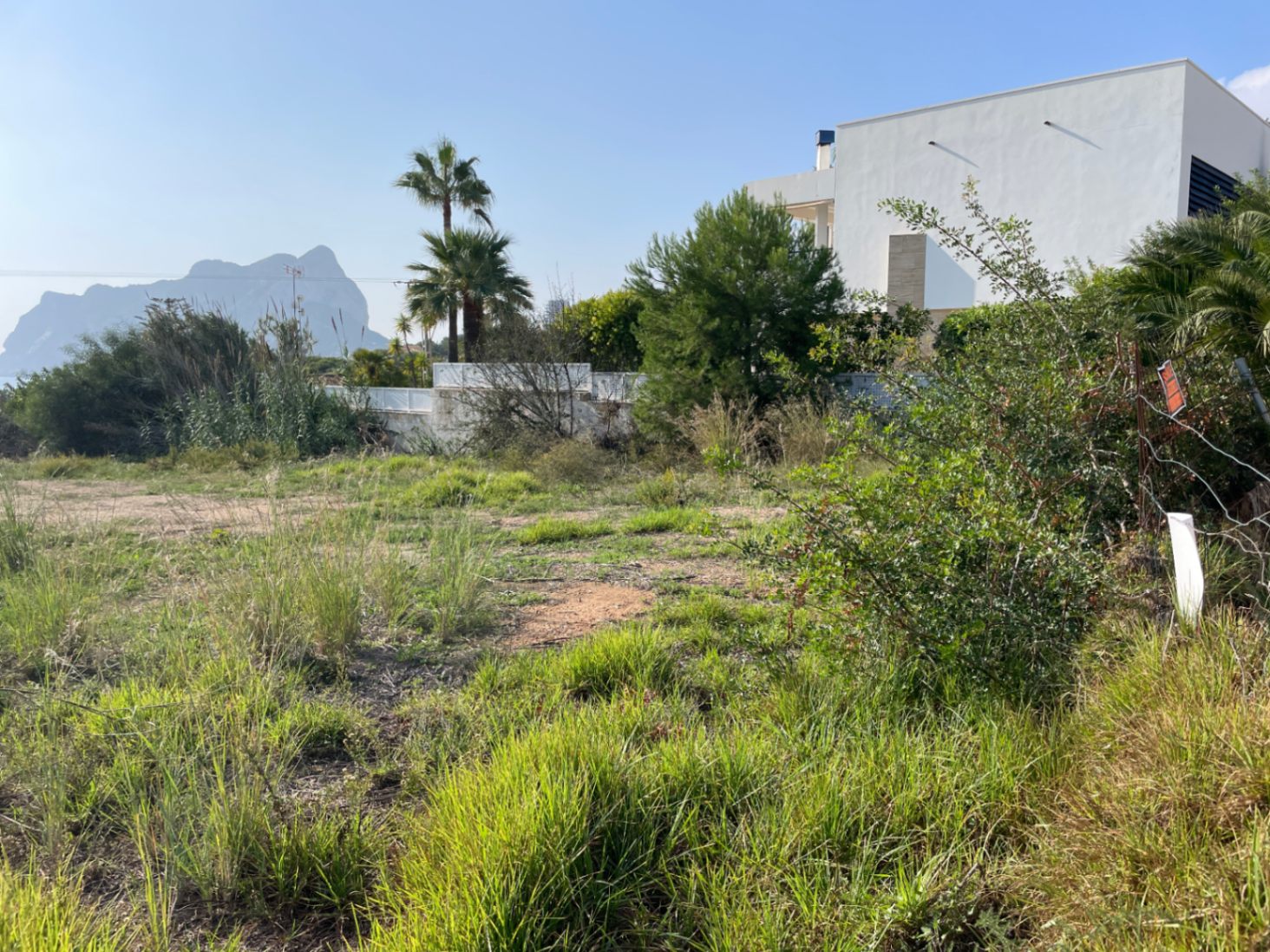 Parcela llana en Bassetes, Calpe, con vistas al Peñón de Ifach. El terreno cuenta con vegetación natural y linda con una villa de arquitectura moderna.