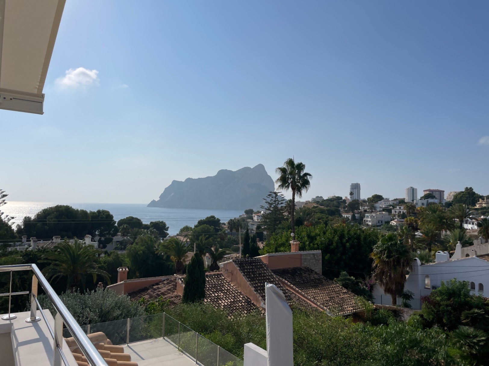 Vista panorámica desde una terraza con barandilla de acero y cristal, mostrando el Peñón de Ifach sobre el mar Mediterráneo y tejados tradicionales.