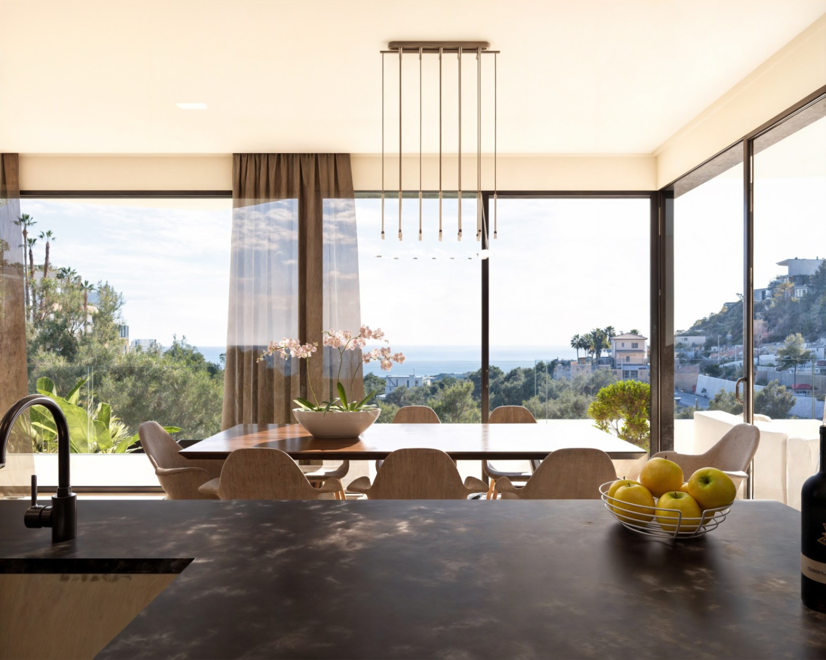 Comedor integrado con mesa de madera y vistas panorámicas al mar a través de grandes ventanales, iluminado por una lámpara lineal de diseño moderno.