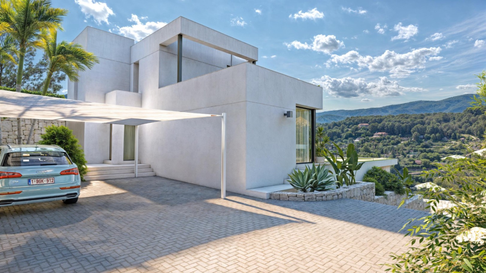 Fachada exterior de diseño cúbico con zona de aparcamiento pavimentada, pérgola textil y muros de piedra natural frente a las vistas de la Sierra de Bernia.