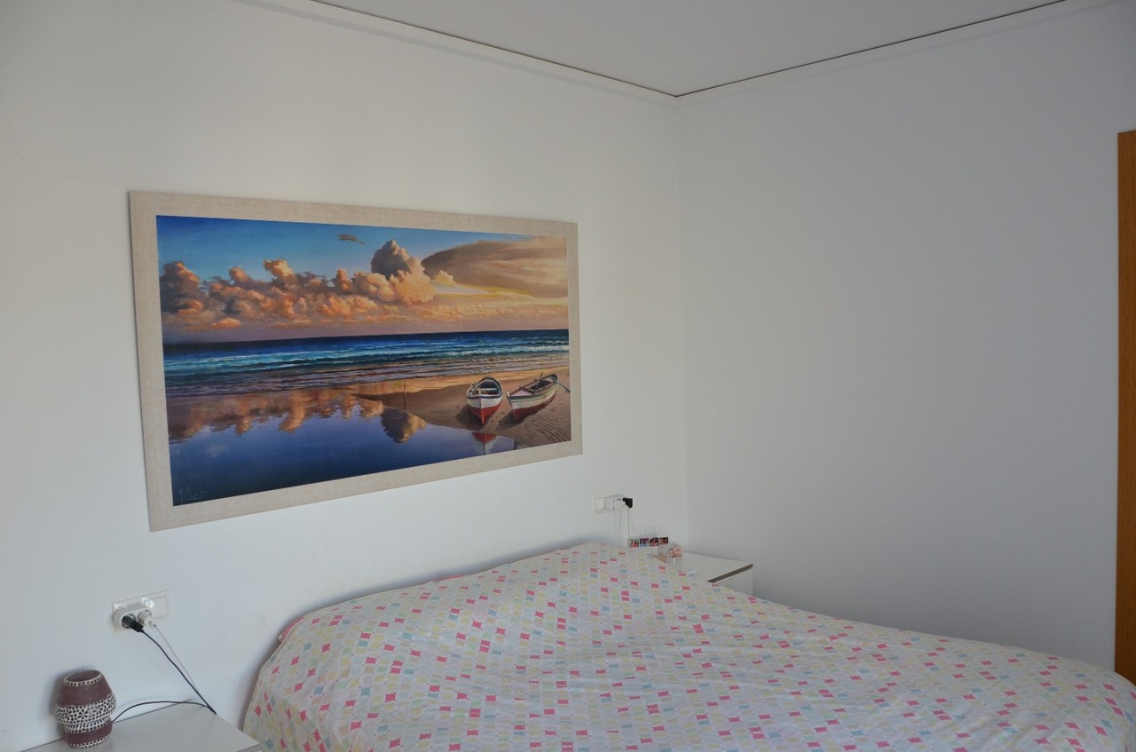 Dormitorio con un gran cuadro de paisaje costero sobre la cama, paredes blancas y una colcha con un patrón geométrico de colores.