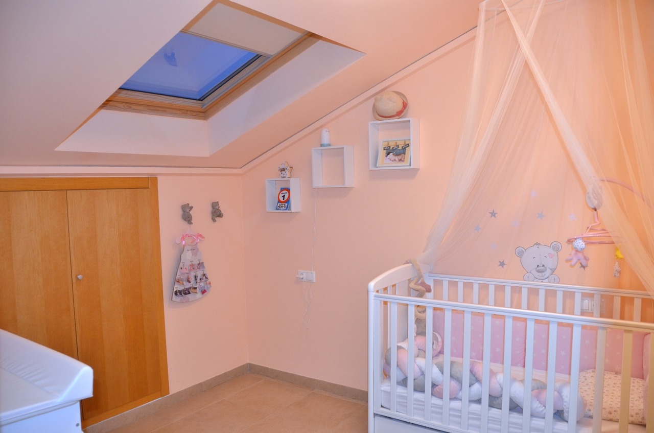 Dormitorio infantil abuhardillado con claraboya, cuna blanca con dosel, paredes en tono rosa y armario empotrado de madera natural.