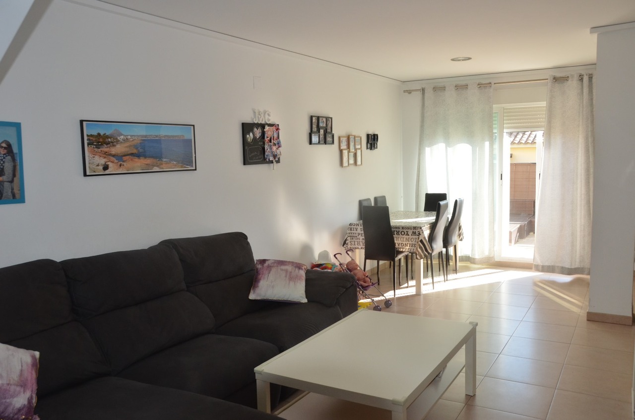 Salón-comedor diáfano con sofá rinconero oscuro, mesa de centro blanca y zona de comedor. La estancia cuenta con suelos cerámicos y acceso directo al exterior mediante ventanales.