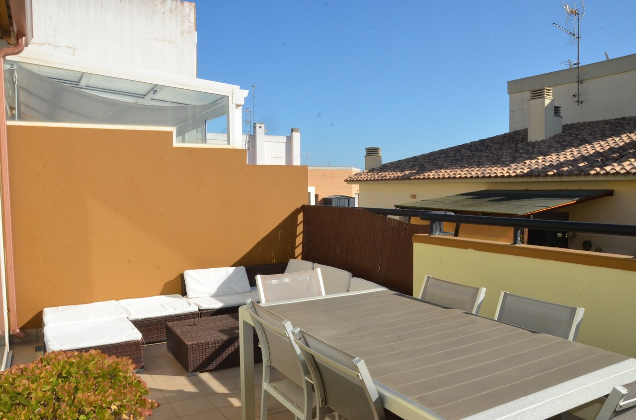 Terraza en la azotea con comedor exterior de aluminio y zona de estar de mimbre con cojines blancos, rodeada de muros en tonos ocre.