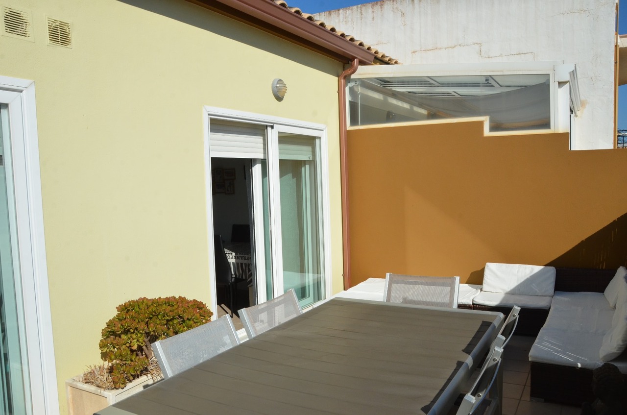 Terraza privada con mesa de comedor exterior, sillas de malla y zona de estar con sofás de cojines blancos junto a un muro perimetral ocre.