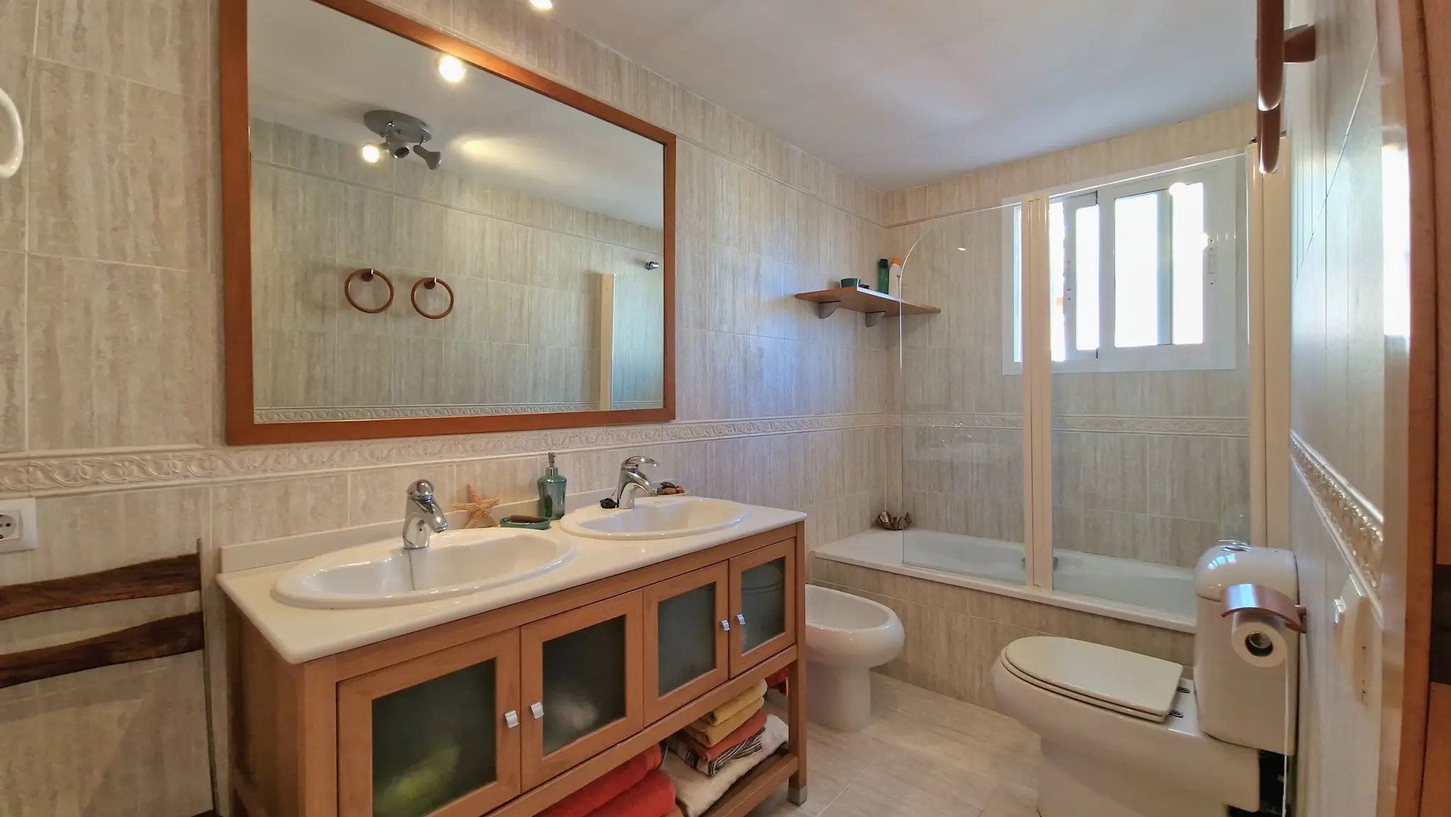 Baño completo con lavabo doble sobre mueble de madera, espejo de gran formato, bidet e inodoro. Incluye bañera con mampara de cristal y paredes alicatadas en tonos crema.