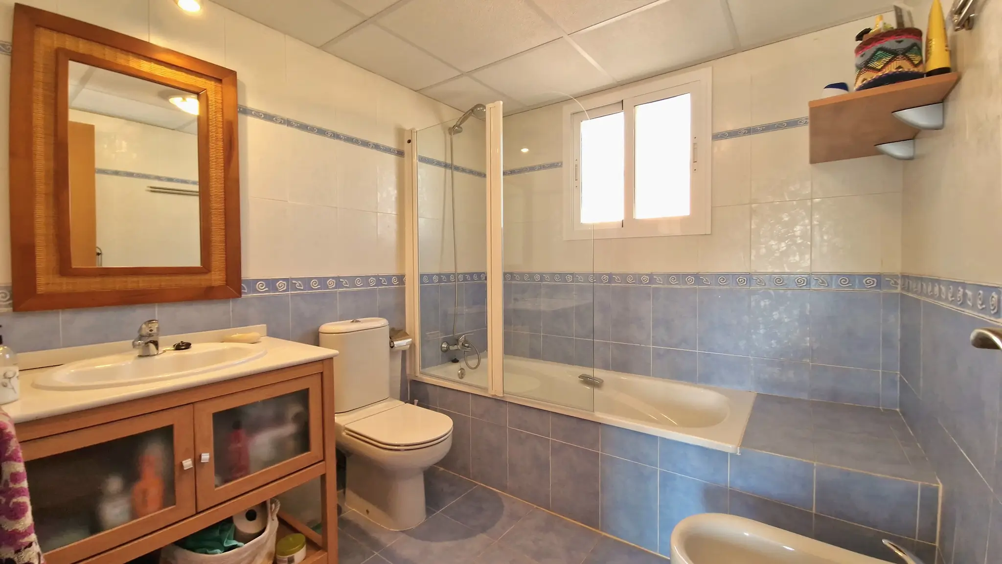 Baño con azulejos azules y blancos, mueble de lavabo en madera y bañera con mampara de cristal, iluminado por luz natural a través de una ventana.