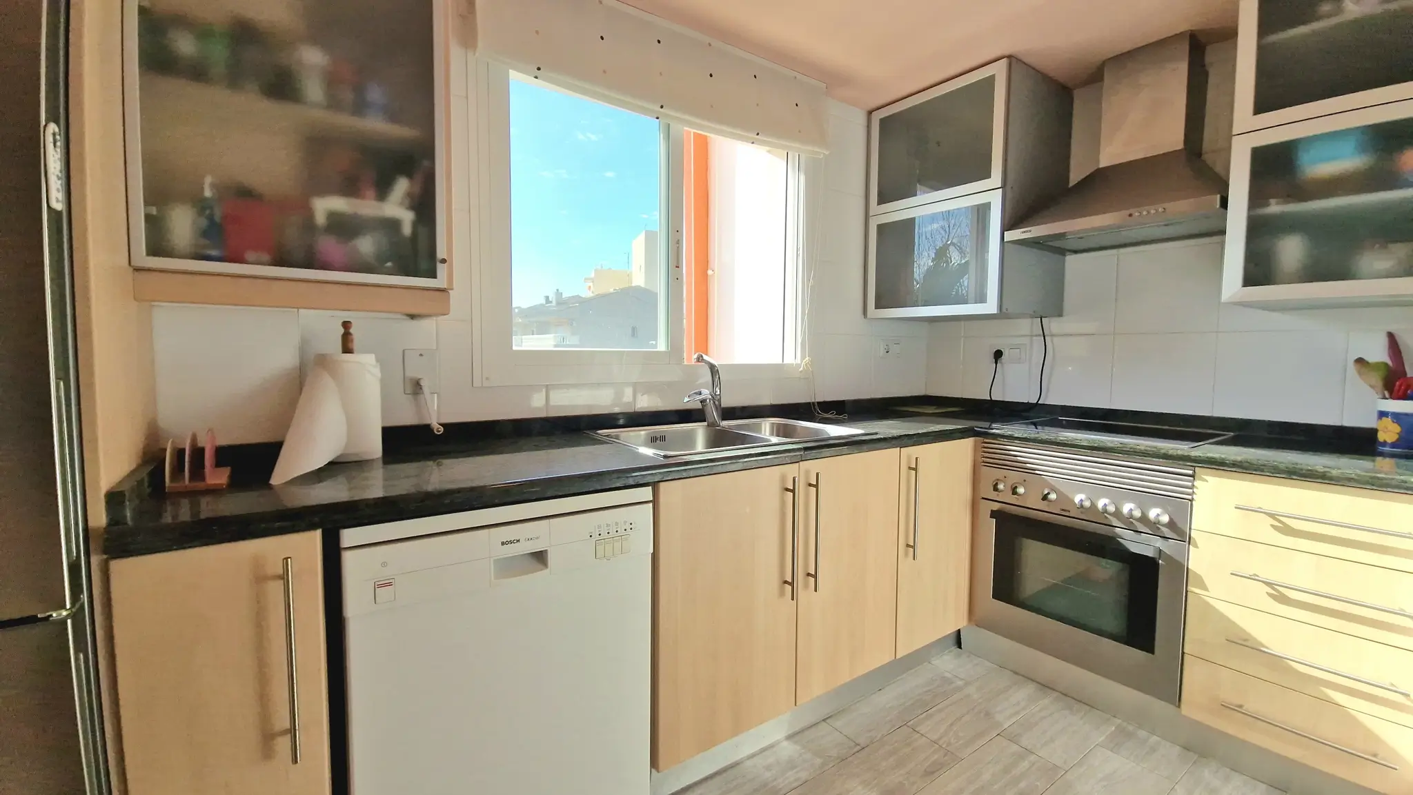 Cocina funcional con armarios de madera clara, encimera de granito oscuro y electrodomésticos integrados, incluyendo lavavajillas Bosch y horno de acero inoxidable.