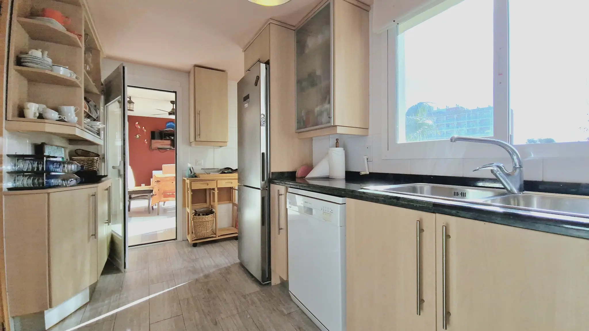 Cocina funcional con mobiliario de madera clara, encimera de granito oscuro, fregadero de acero inoxidable y acceso directo al salón comedor.
