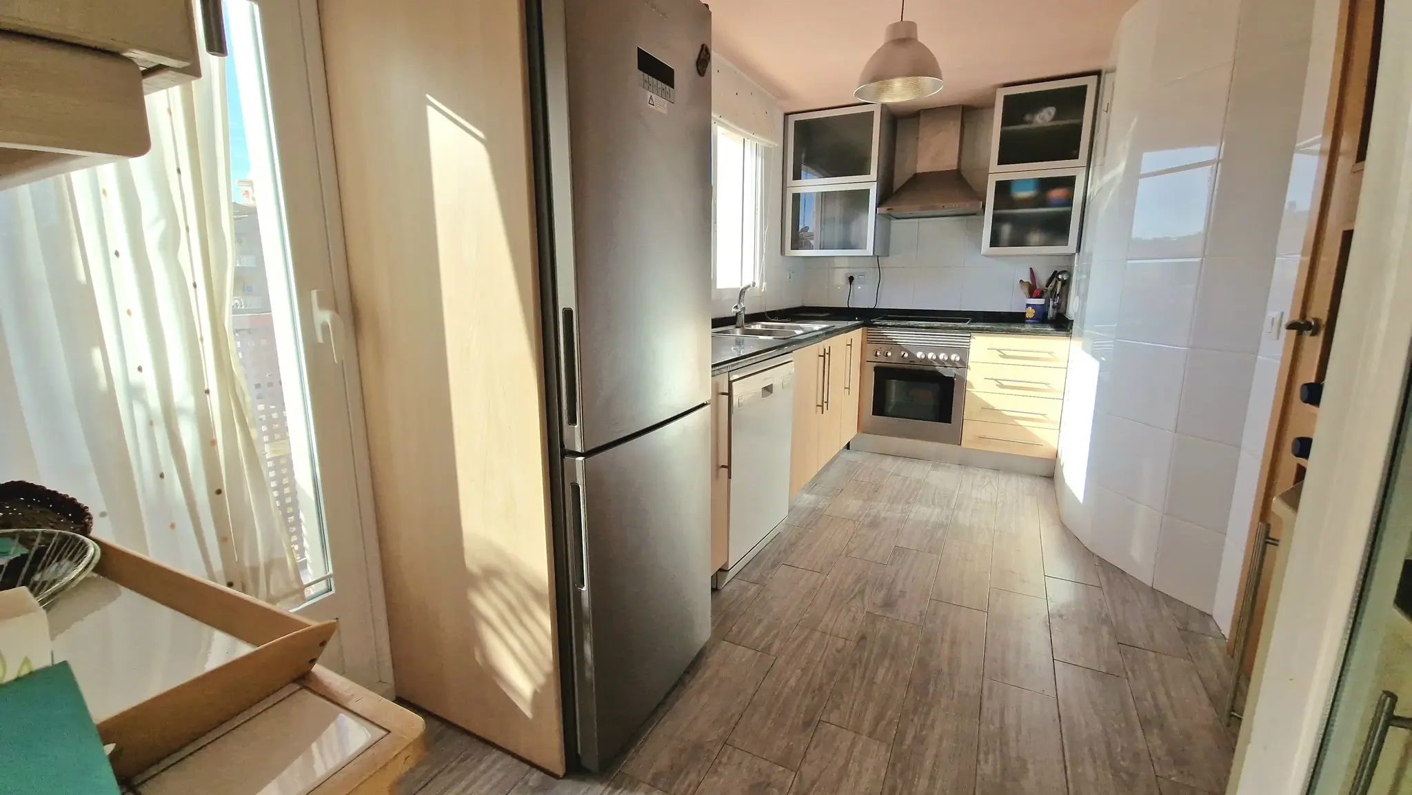 Cocina moderna con electrodomésticos de acero inoxidable, armarios de madera clara y suelo de gres cerámico efecto madera iluminado por luz natural.