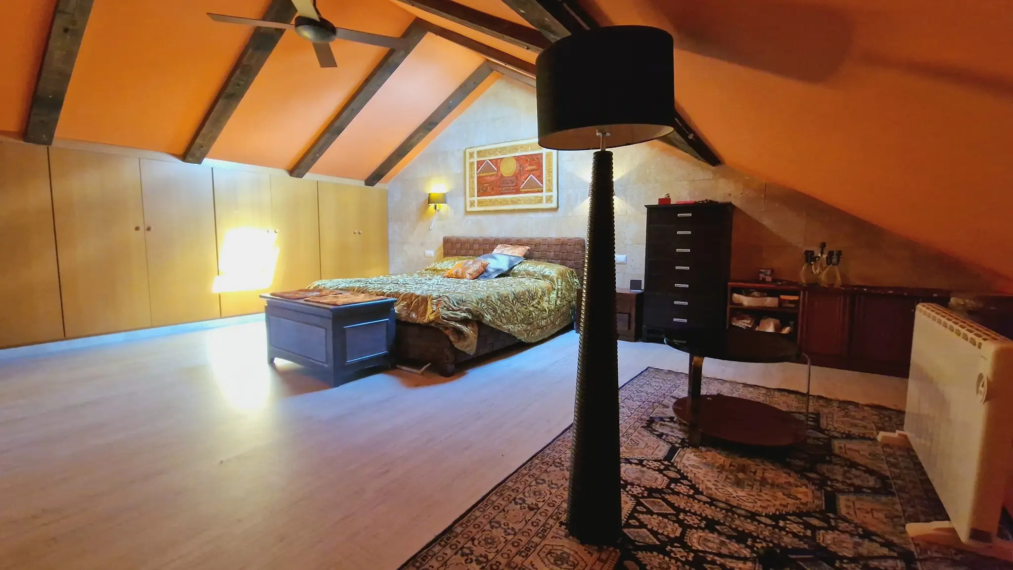 Dormitorio abuhardillado con vigas de madera oscura, armarios empotrados de madera clara y una pared frontal de piedra natural bajo el techo inclinado.