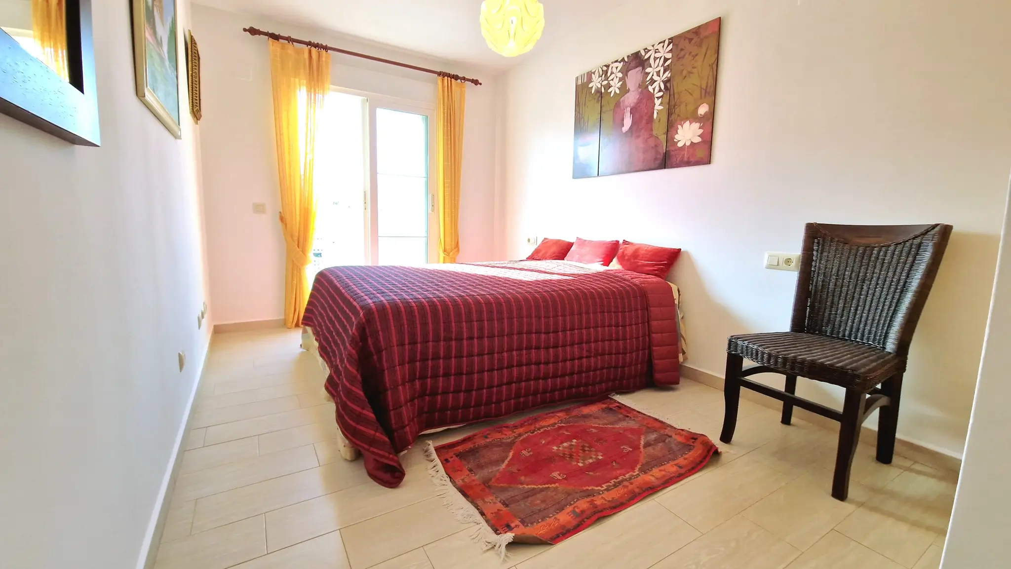 Dormitorio luminoso con cama doble, colcha roja acolchada, cortinas amarillas, silla de mimbre oscura y un tríptico decorativo de Buda en la pared.