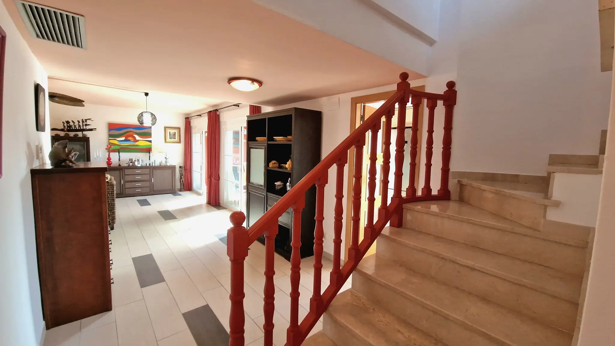 Escalera de mármol con barandilla de madera roja que conecta con un salón luminoso de suelos cerámicos y mobiliario de madera oscura.