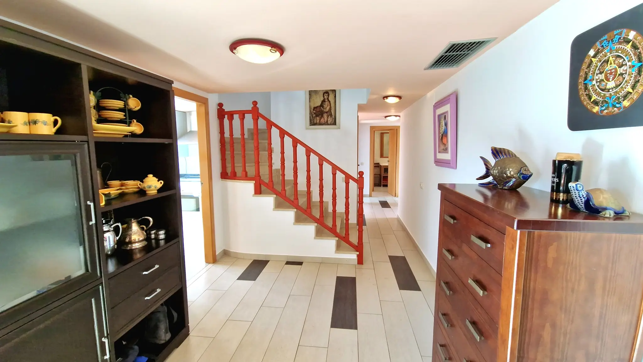 Pasillo con escalera de barandilla roja de madera, suelos cerámicos con detalles oscuros, vitrina de madera oscura y cómoda lateral con elementos decorativos.