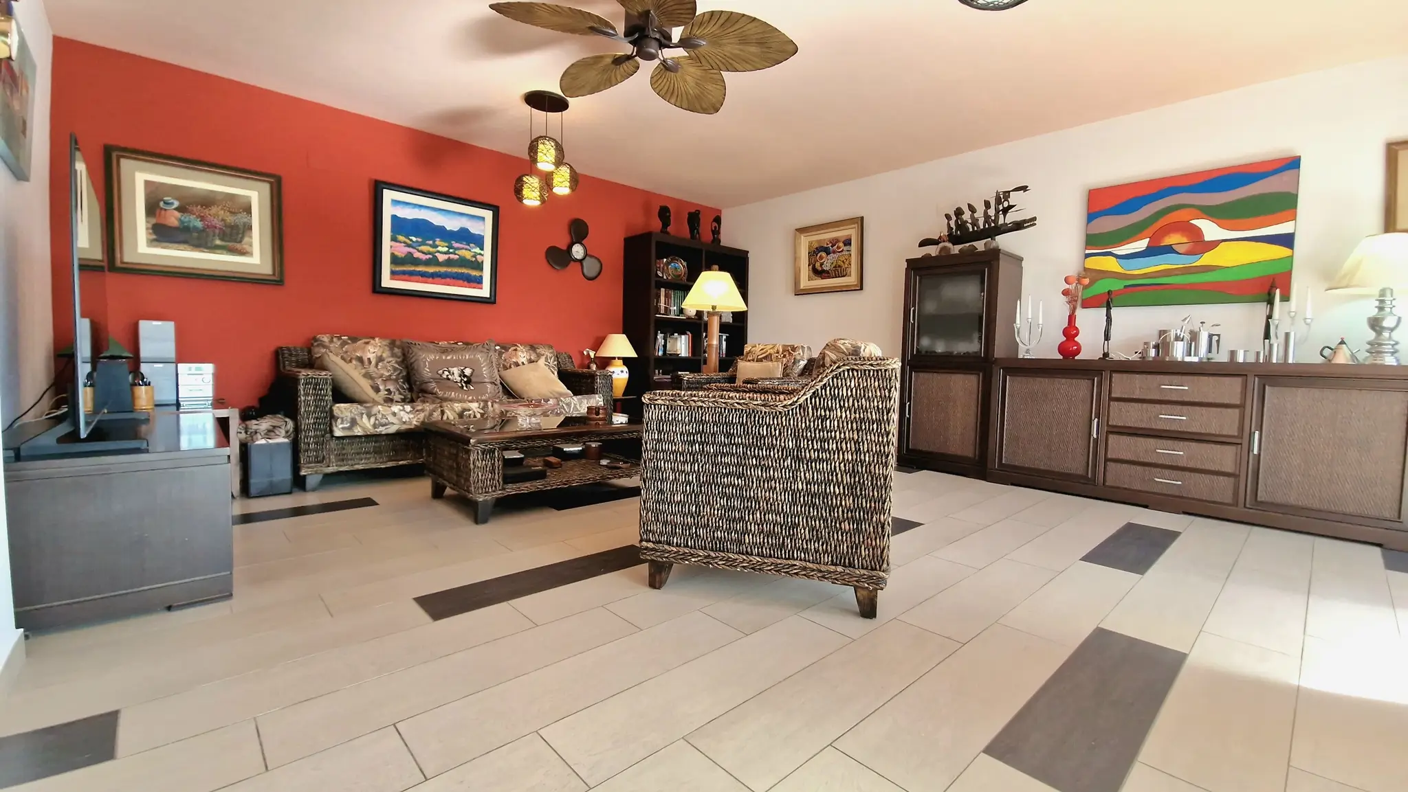 Salón con mobiliario de mimbre, pared de acento terracota con arte enmarcado y ventilador de techo con aspas de diseño tropical sobre suelo cerámico.