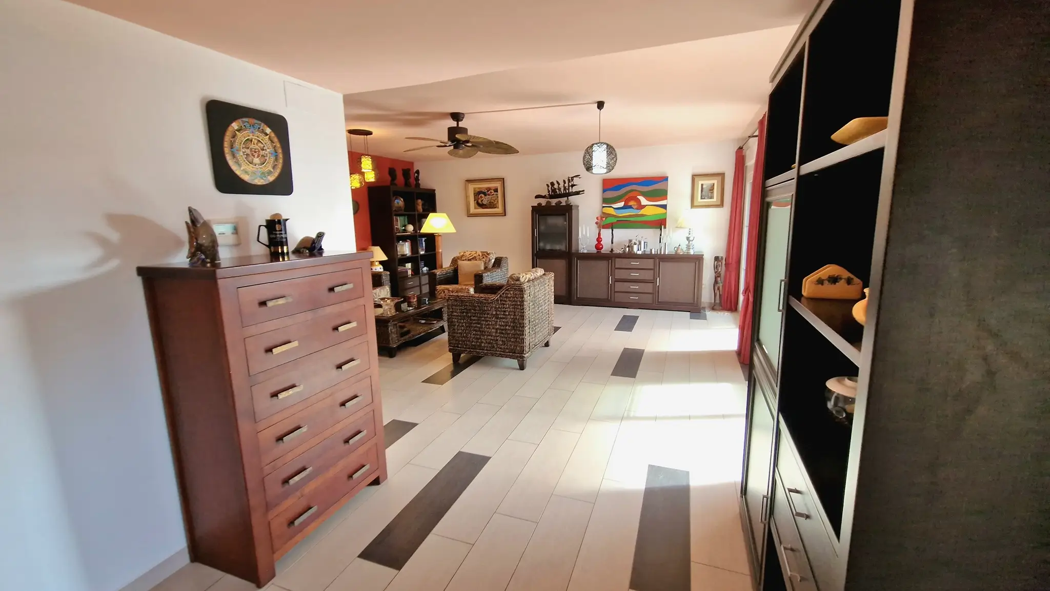 Salón espacioso con suelo cerámico de tonos claros y oscuros, mobiliario de mimbre, una cómoda de madera y ventilador de techo integrado.