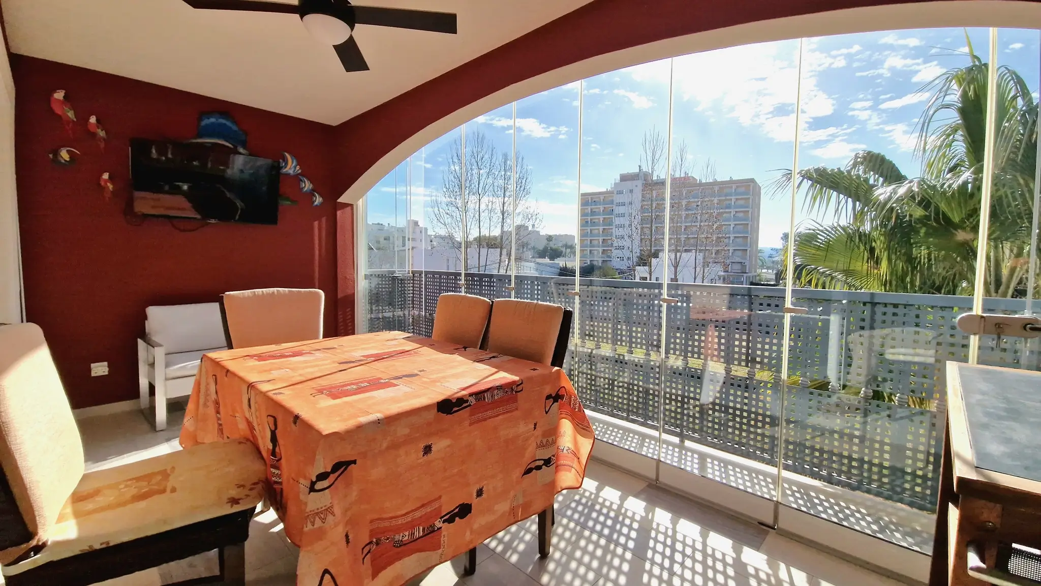 Terraza acristalada con cortinas de cristal, zona de comedor, pared de acento roja con televisor y vistas despejadas al entorno urbano.