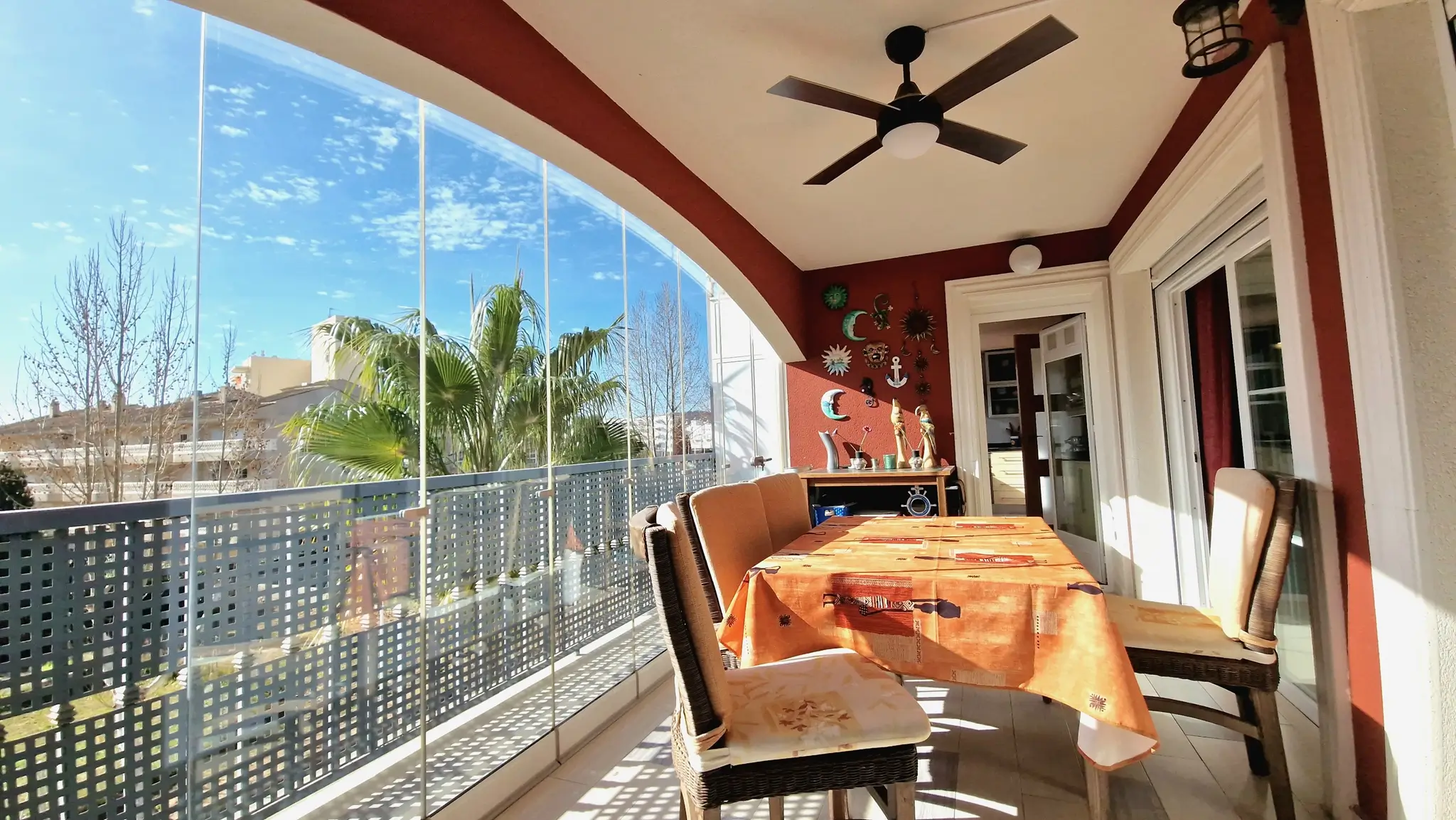 Terraza cubierta con cerramiento de cristal sin perfiles, comedor exterior con sillas de mimbre, ventilador de techo y vistas despejadas a palmeras.