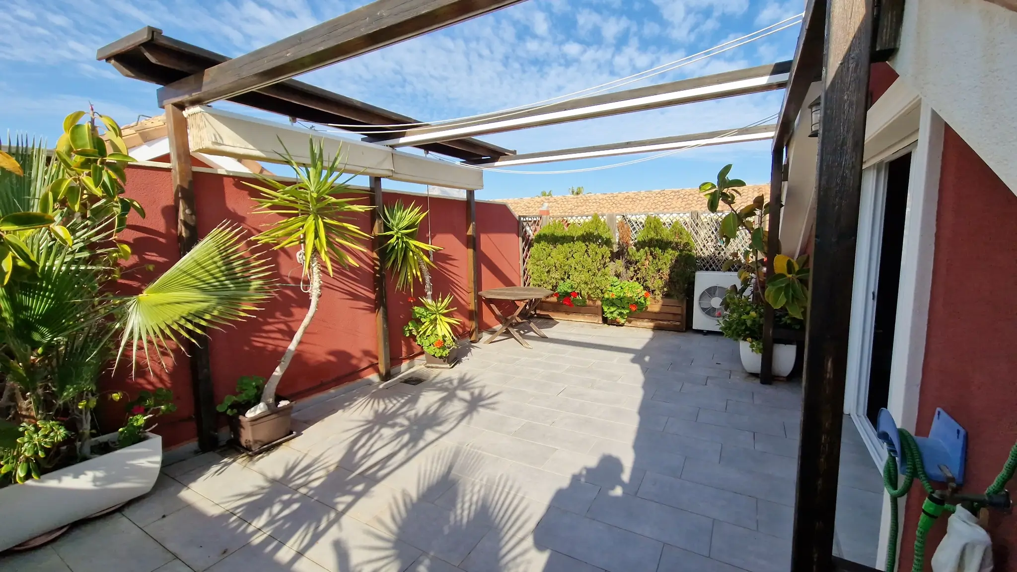 Terraza privada con suelo de baldosas grises, pérgola de madera oscura y toldos blancos. Destacan las paredes color teja, plantas ornamentales y una mesa de madera.