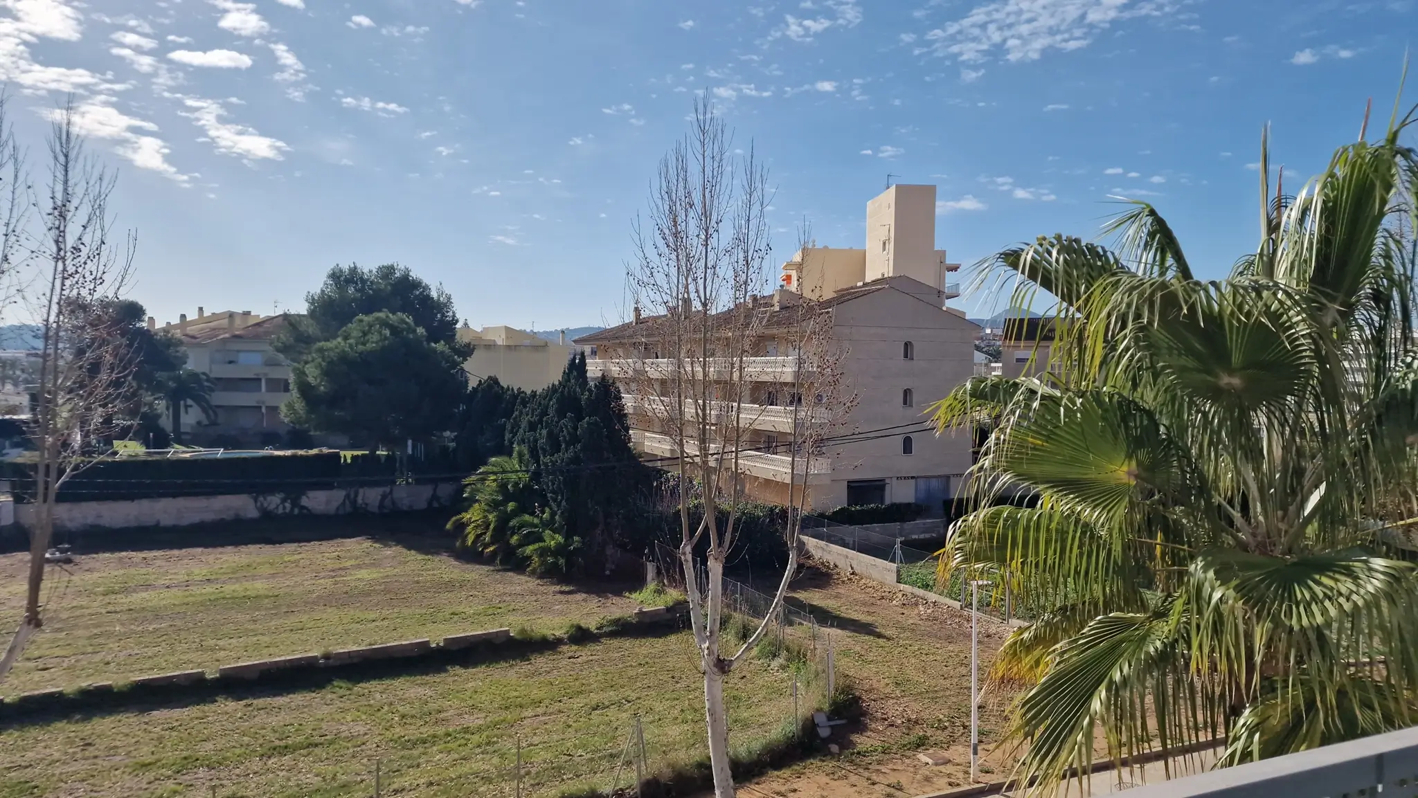 Vista exterior elevada hacia una parcela despejada y edificios residenciales, enmarcada por vegetación mediterránea y árboles caducifolios bajo un cielo despejado.