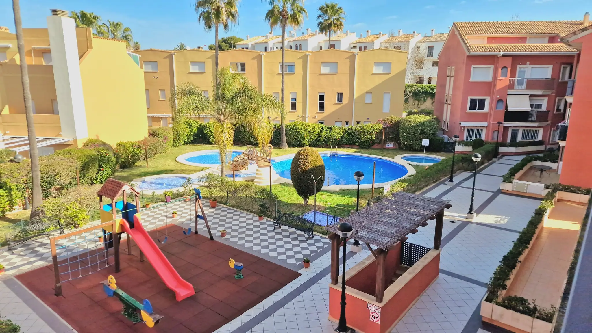 Zona comunitaria con parque infantil, piscina de diseño con puente y jardines cuidados, rodeada de edificios residenciales de estilo mediterráneo bajo un cielo despejado.