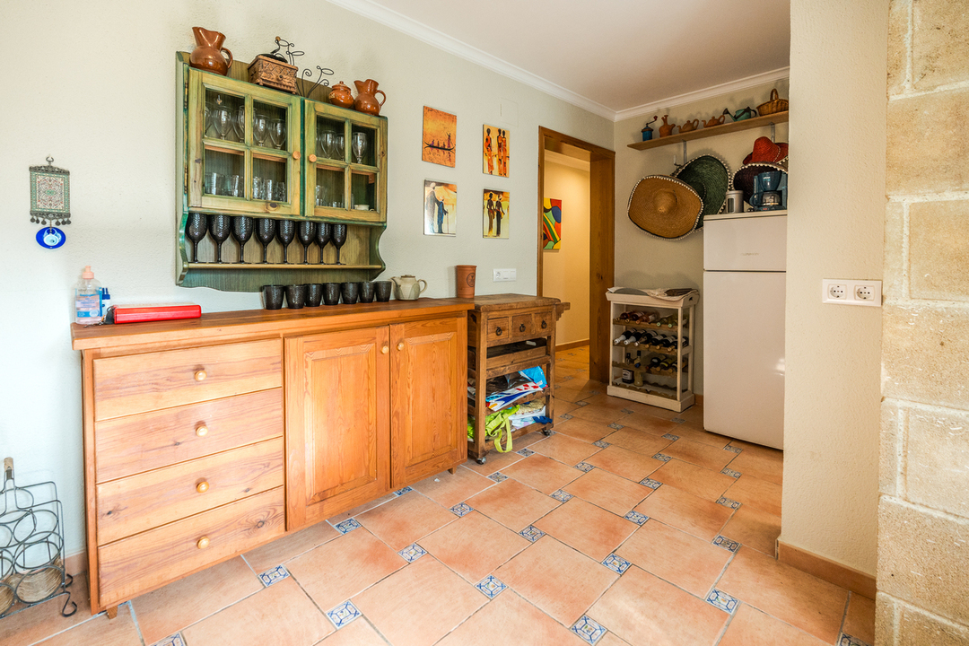 Aparador de madera rústica y vitrina verde con cristalería sobre suelo de terracota con detalles decorativos. Incluye un botellero y nevera blanca en un ambiente tradicional.