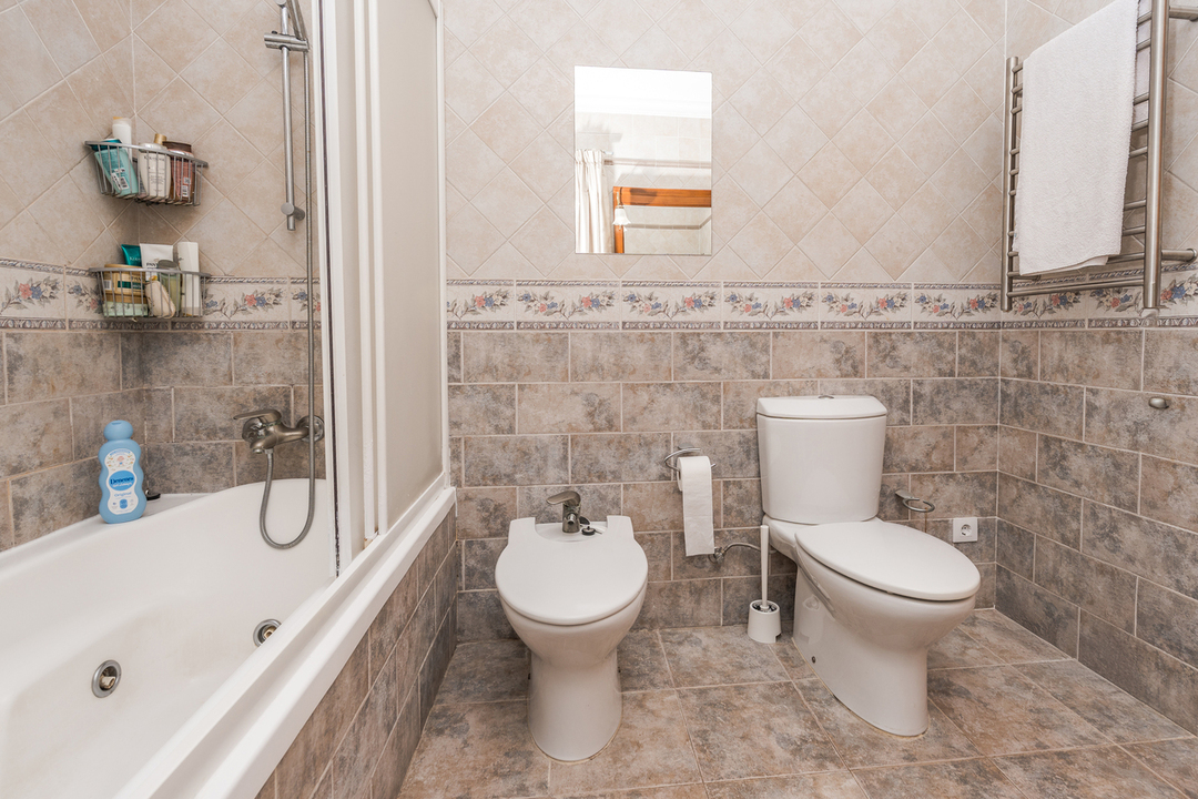 Baño completo con bañera, bidé e inodoro, revestido con azulejos en tonos tierra y una cenefa floral decorativa. Incluye un radiador toallero de acero inoxidable.