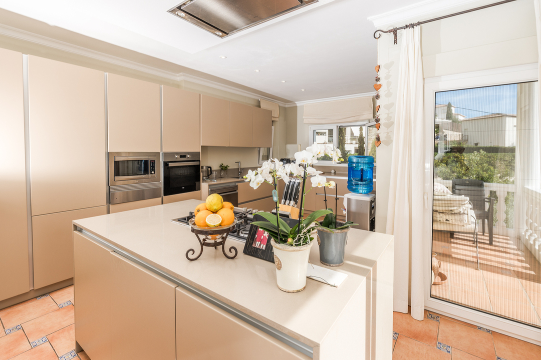Cocina moderna con isla central beige, placa de gas, electrodomésticos integrados y suelos de terracota. Puertas acristaladas conectan directamente con la terraza exterior.